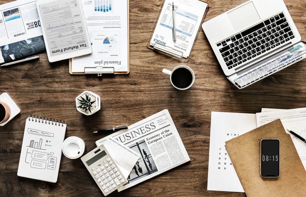 Analisis Ekonomi Mikro: Deskriptif, Normatif, dan Positif 1 Tampilan dari atas meja kayu dengan berbagai dokumen keuangan, laptop, kalkulator, ponsel pintar, cangkir kopi, dan tanaman kecil.
